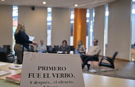 Mujer con cabello rubio oscuro habla en aula, mientras cuatro personas escuchan atentamente. Libro sobre mesa.