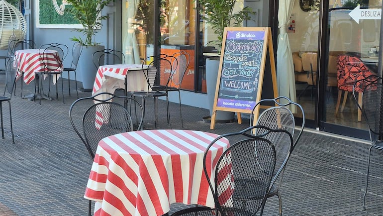 Ambiente exterior de café con mesas y sillas vacías. Cartel de bienvenida en pizarra. Espacio iluminado y decorado con plantas.