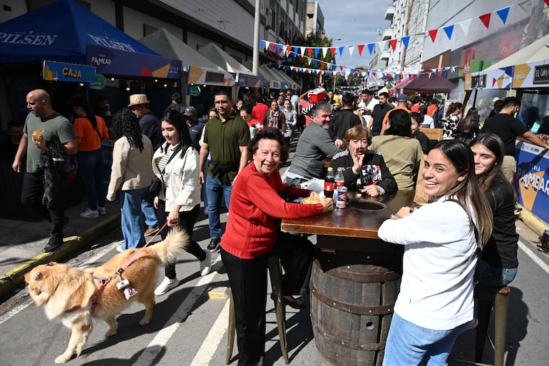 Cuatro mujeres sonríen en una mesa alta, rodeadas de decoraciones festivas en una feria popular con stands de comida.