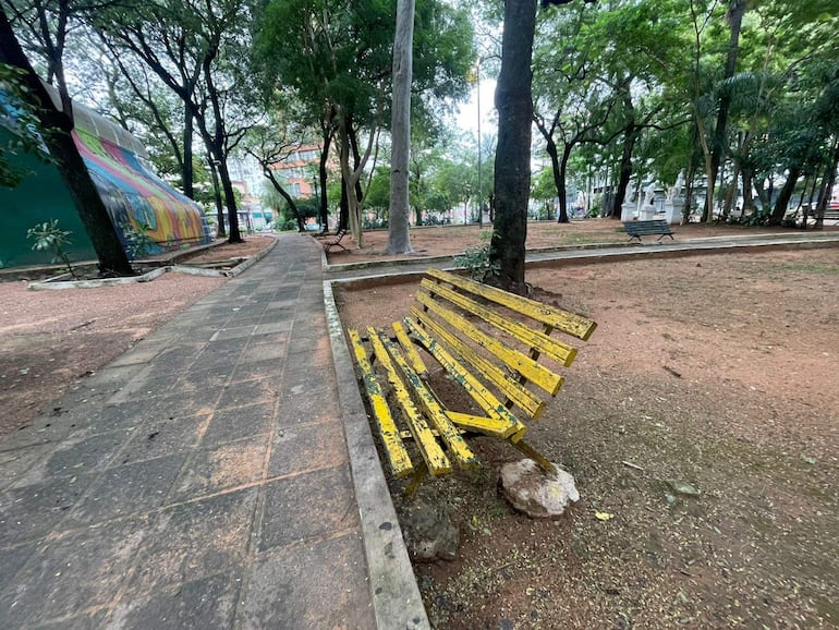 Banco amarillo deteriorado inclinado sobre una roca en un parque vacío, rodeado de áreas verdes y un mural colorido.