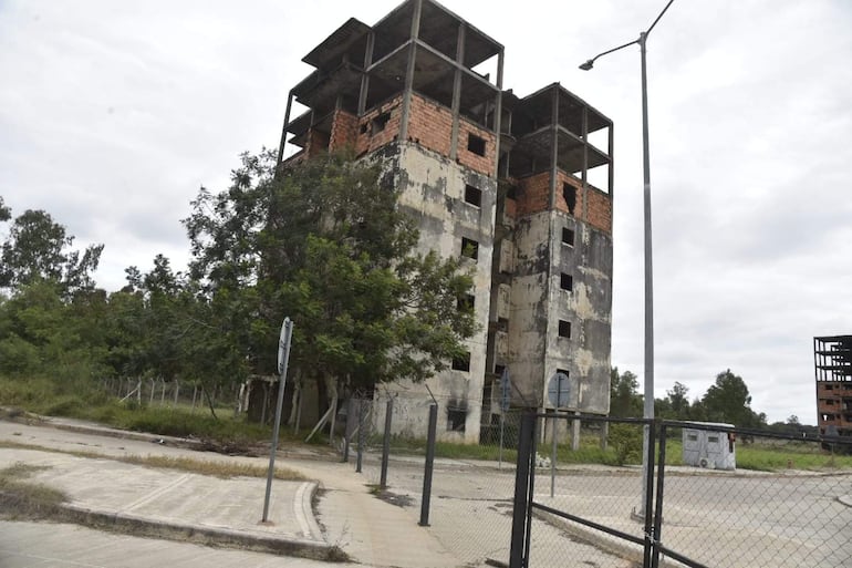 Construcción inacabada en estado de abandono, rodeada de vegetación y pavimento deteriorado, bajo un cielo nublado.
