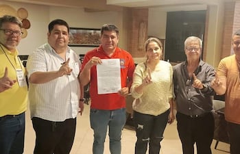 Seis personas en un ambiente moderno levantan el dedo índice, sonriendo y sosteniendo documentos. Visten camisetas de diversos colores.