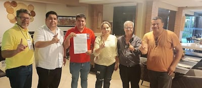 Seis personas en un ambiente moderno levantan el dedo índice, sonriendo y sosteniendo documentos. Visten camisetas de diversos colores.