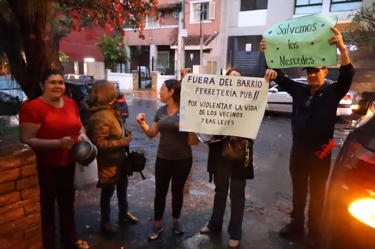 Grupo de cuatro personas, una con olla y otra con abrigo, sostienen carteles pidiendo el cierre del bar en un ambiente nocturno.