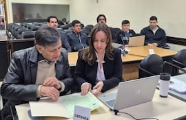 Gómez con chaqueta de cuero y defensora en blazer, revisan documentos en una sala de tribunal con público atento.