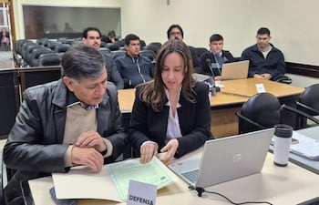 Gómez con chaqueta de cuero y defensora en blazer, revisan documentos en una sala de tribunal con público atento.