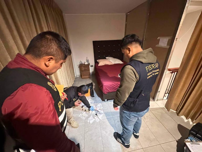 Dos oficiales de policía con chalecos distintivos observan a un hombre agachado analizando objetos en el suelo en una habitación oscura.