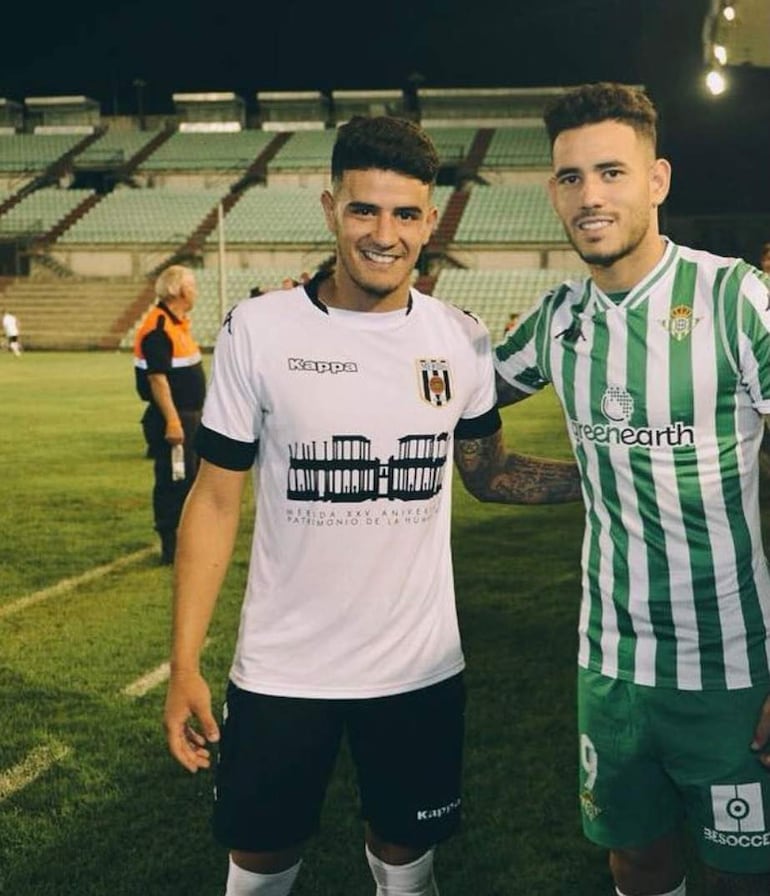 Antonio Sanabria y otro jugador sonríen, ambos con camisetas de fútbol. Fondo de gradas vacías.