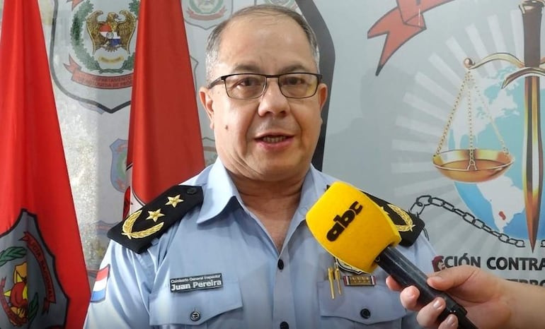 Juan Pereira, con cabello corto y gafas, en uniforme celeste claro, sostiene un micrófono amarillo mientras habla en un evento oficial, rodeado de banderas rojas.