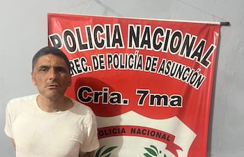 Hombre con camiseta blanca y tatuaje visible, de pie frente a un cartel de la Policía Nacional en un ambiente interior.