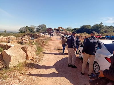 Seis policías en camino de tierra, uno hablando con otro cerca de un vehículo de la policía, excavadora trabajando al fondo.