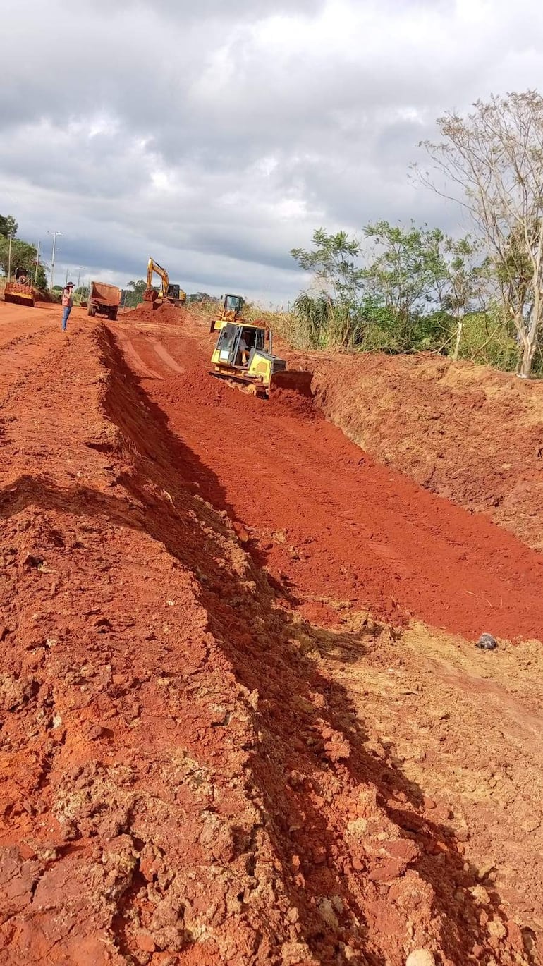 Hombre observa de pie en un terreno rojo, rodeado de maquinaria pesada como excavadoras en un ambiente nublado y rural.