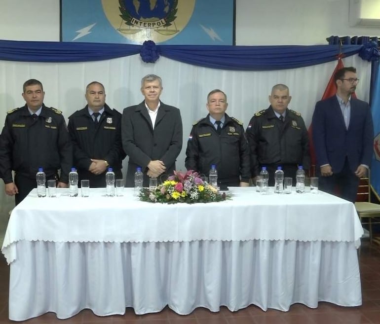 Hombres en uniforme de policía y traje oscuro en evento con logo de Interpol y banderas de fondo, rodeados de botellas y un arreglo floral.