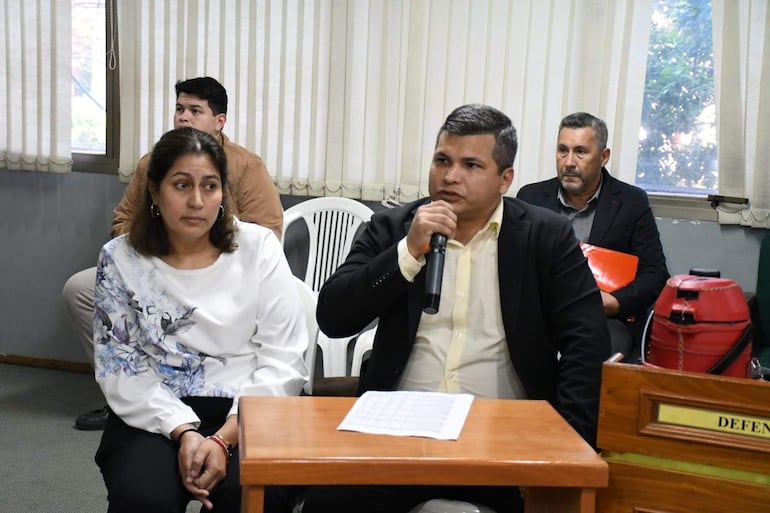 José Jara con micrófono, mujer con camiseta blanca y estampados florales escuchando, hombres en fondo en reunión formal.
