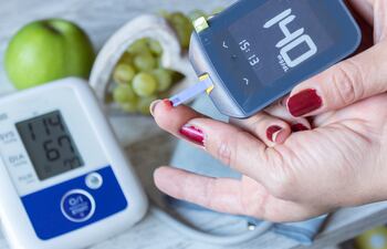 Una mujer con las uñas pintadas de rojo y descascaradas se mide la glucosa con un pinchazo en el dedo y el resultado es de 140. Sobre la mesa, un aparato medidor de la presión arterial muestra que tiene 114/67. También hay una manzana verde y un racimo de uvas.