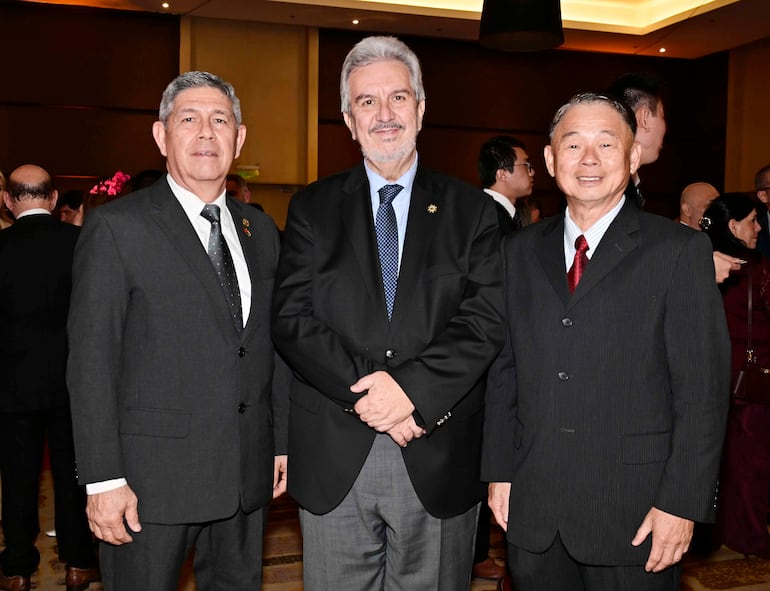Tres hombres con trajes: uno en corbata negra, otro en camisa azul y saco gris, y uno más con corbata roja, sonrientes en un evento.
