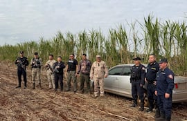 Ocho personas armadas alineadas en zona rural, junto a un automóvil blanco desatendido, rodeadas de caña de azúcar.