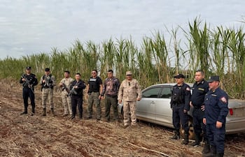Ocho personas armadas alineadas en zona rural, junto a un automóvil blanco desatendido, rodeadas de caña de azúcar.