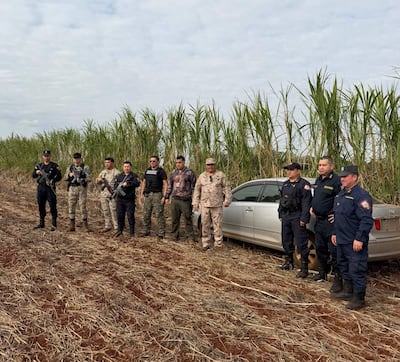 Ocho personas armadas alineadas en zona rural, junto a un automóvil blanco desatendido, rodeadas de caña de azúcar.