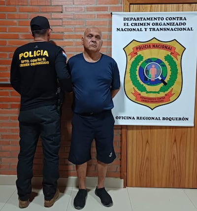 Hombre canoso con camiseta azul y pantalones oscuros de pie con brazos cruzados, junto a un policía en uniforme negro en oficina institucional.