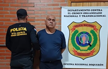 Hombre canoso con camiseta azul y pantalones oscuros de pie con brazos cruzados, junto a un policía en uniforme negro en oficina institucional.