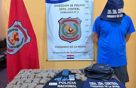 Hombre con capuchón azul frente a mesa con paquetes de drogas y dinero en efectivo, con bandera policial al lado.