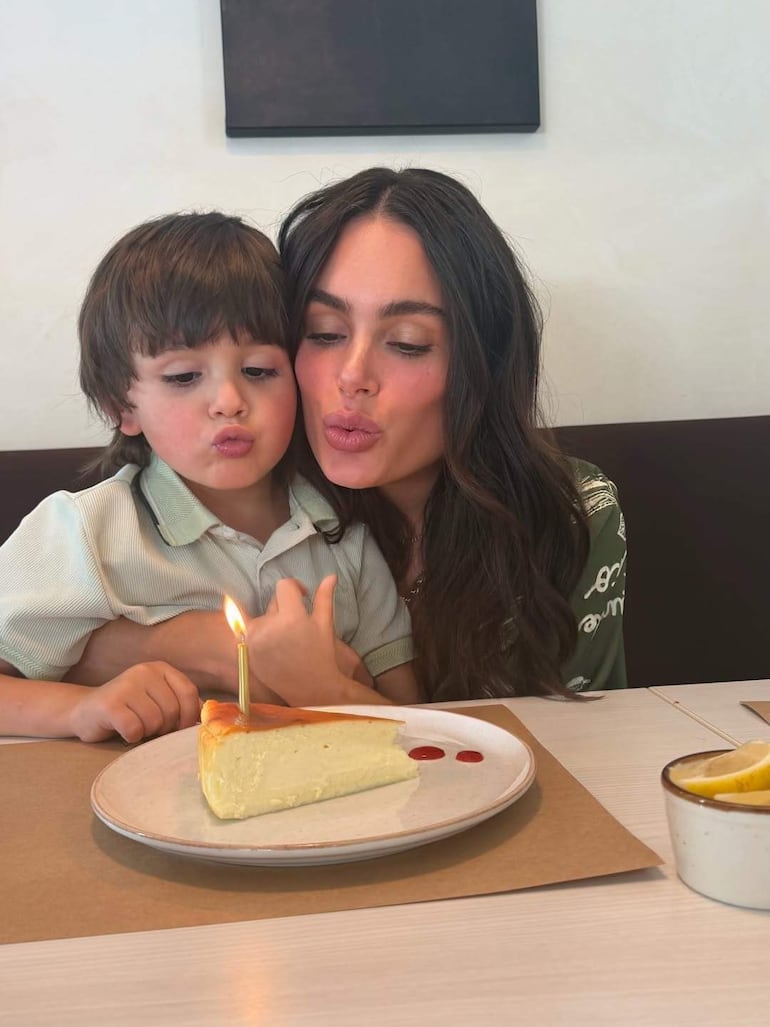 Niño a la izquierda y mujer a la derecha sonríen mientras posan junto a un pastel con vela encendida en un ambiente acogedor.