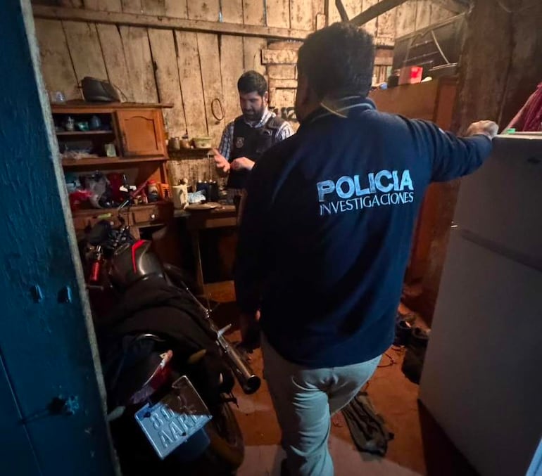 Hombre con chaleco hablando frente a otro con abrigo marcado como Policía Investigaciones en un ambiente interior desordenado.