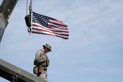 Soldado de pie con uniforme de camuflaje y chaleco táctico, mirando hacia abajo, con bandera de EE.UU. ondeando.