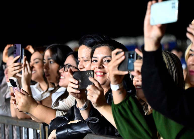 Seis mujeres sonrientes en primer plano, grabando con sus teléfonos móviles en un ambiente iluminado de concierto.