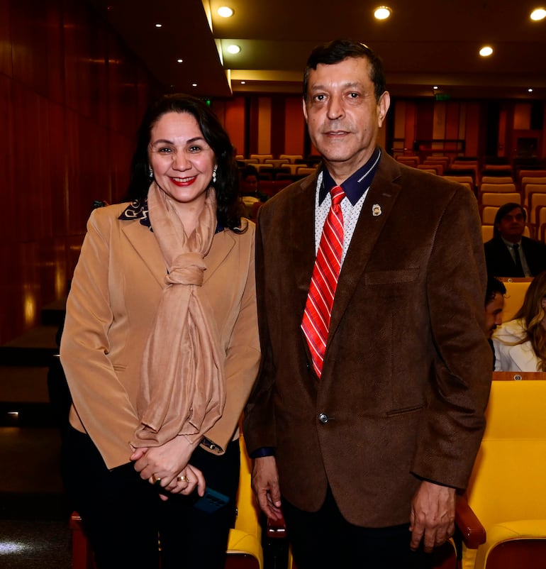 Mujer con abrigo claro y bufanda sonríe, junto a hombre en blazer marrón y corbata roja en un evento formal.
