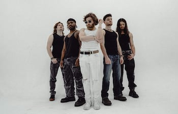 Cinco hombres de pie en ambiente minimalista. Varón de cabello rizado en camiseta negra, junto a otros con diferentes vestimentas oscuras.