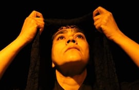 Mujer en primer plano con velo oscuro, mirada concentrada y manos levantando el velo en un ambiente teatral iluminado.