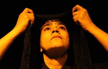 Mujer en primer plano con velo oscuro, mirada concentrada y manos levantando el velo en un ambiente teatral iluminado.