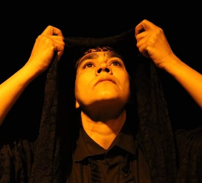 Mujer en primer plano con velo oscuro, mirada concentrada y manos levantando el velo en un ambiente teatral iluminado.