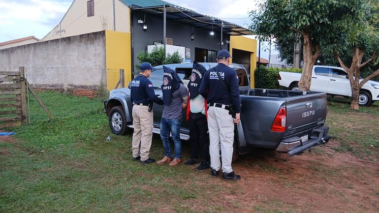 Cuatro agentes de policía en uniforme sostienen un hombre con capucha en un área verde, junto a una vivienda clara y una camioneta gris.