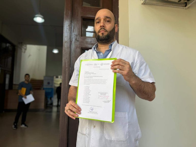 Hombre en delantal blanco sostiene un documento frente a un edificio institucional, con expresión seria.
