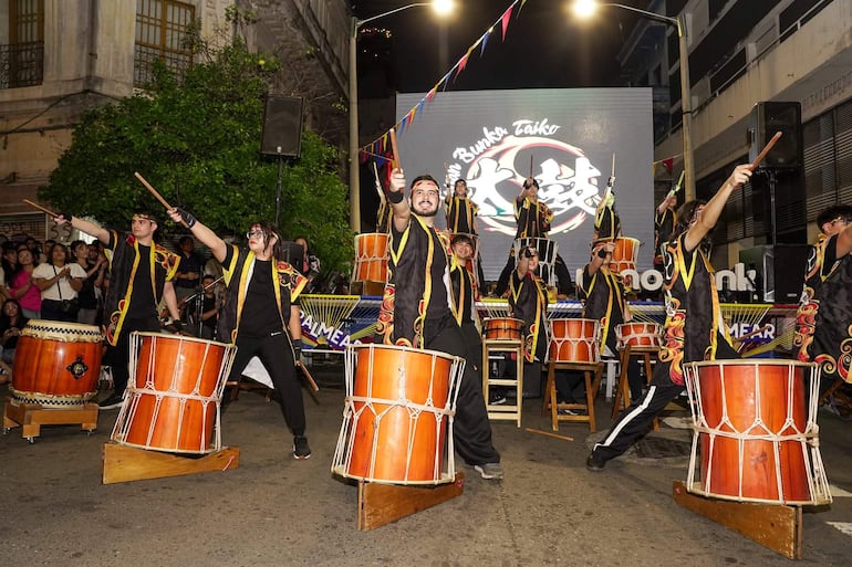 Grupo de personas vestidas de negro con detalles coloridos, tocando tambores en un ambiente festivo con decoraciones y luces.