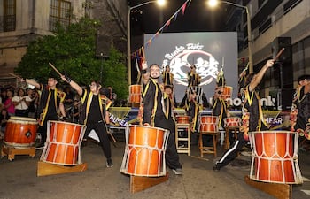 Grupo de personas vestidas de negro con detalles coloridos, tocando tambores en un ambiente festivo con decoraciones y luces.