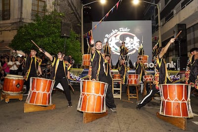 Grupo de personas vestidas de negro con detalles coloridos, tocando tambores en un ambiente festivo con decoraciones y luces.