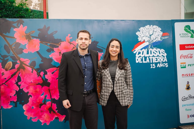 Hombre con barba y camisa azul junto a mujer en blusa gris, ambos sonríen en fondo decorativo de flores.