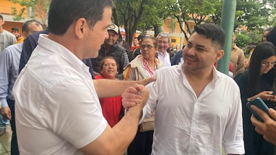 Dos hombres de pie sonriendo y dándose la mano, uno con camisa blanca, rodeados de personas en un ambiente al aire libre.