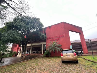 Hospital Regional de Caacupé, edificio rojo y moderno con vehículos estacionados y árboles alrededor, en un día nublado.