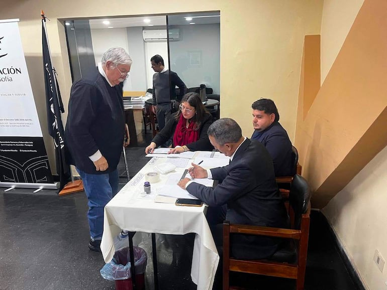 Hombre mayor con suéter negro dirige la sala, mientras mujer con bufanda roja escribe y otros observan en un ambiente formal.