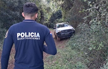 Hombre vestido con camiseta azul marino de 'POLICÍA INVESTIGACIONES' camina por sendero, mientras observa una camioneta blanca parcialmente cubierta de barro.
