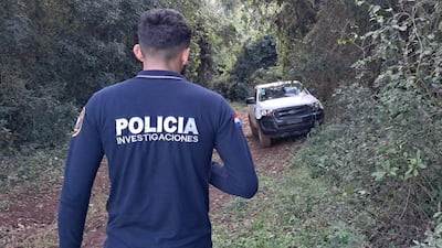 Hombre vestido con camiseta azul marino de 'POLICÍA INVESTIGACIONES' camina por sendero, mientras observa una camioneta blanca parcialmente cubierta de barro.