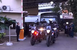 Seis agentes de seguridad en motocicletas, con atuendos oscuros y cascos, resguardan una camioneta Mercedes-Benz frente a un edificio.