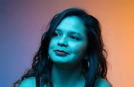 Mujer de cabello rizado y camiseta negra observa con cámara en mano, rodeada de un ambiente iluminado en tonos azules y anaranjados.