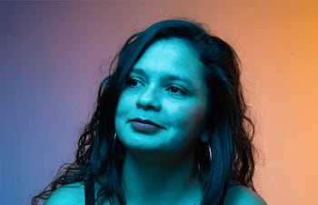 Mujer de cabello rizado y camiseta negra observa con cámara en mano, rodeada de un ambiente iluminado en tonos azules y anaranjados.