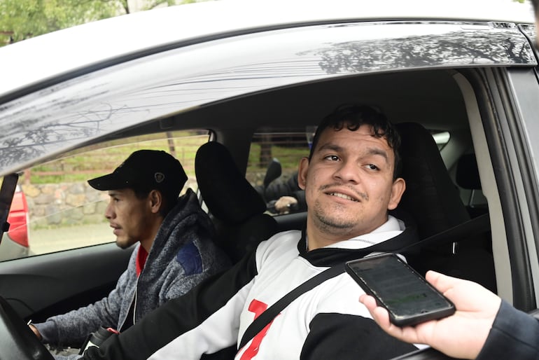 Dos hombres, uno con camiseta blanca y otro con abrigo gris, dentro de un auto, sosteniendo un teléfono móvil en un entorno urbano.